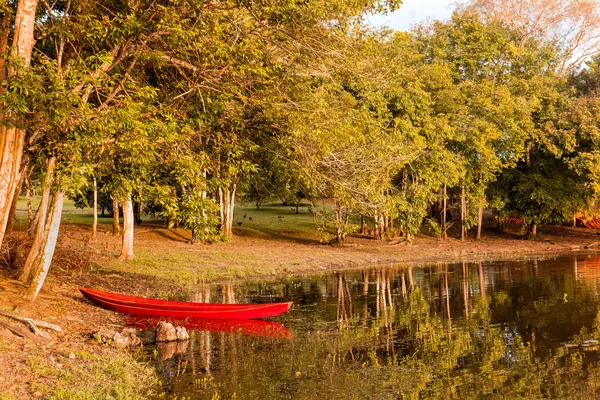 Uma canoa vermelha à beira d'água entre árvores em tons quentes.