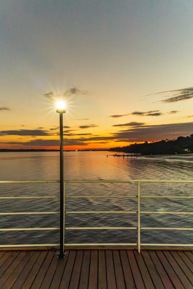 Um poste aceso no deck à beira do rio enquanto o sol se põe no horizonte.