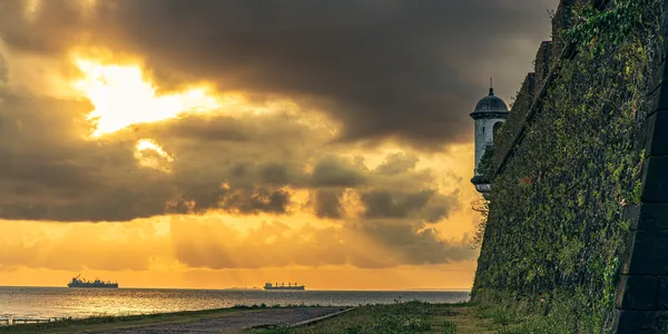 O sol rompe as nuvens sobre o rio ao lado da muralha e da guarita da Fortaleza.