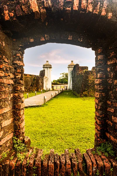 Uma abertura em arco no tijolo antigo enquadra o gramado e as guaritas da Fortaleza.