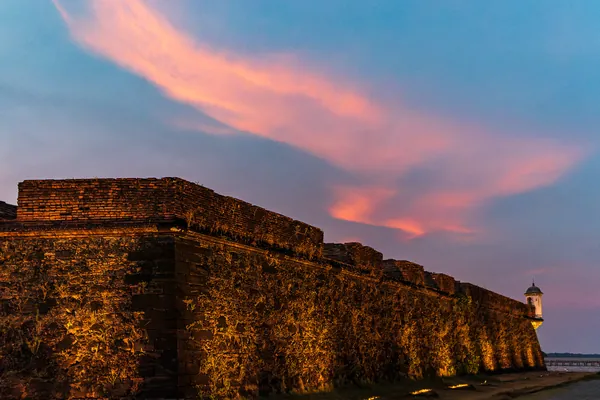 A muralha de pedra se estende sob nuvens rosadas e céu azul ao entardecer.