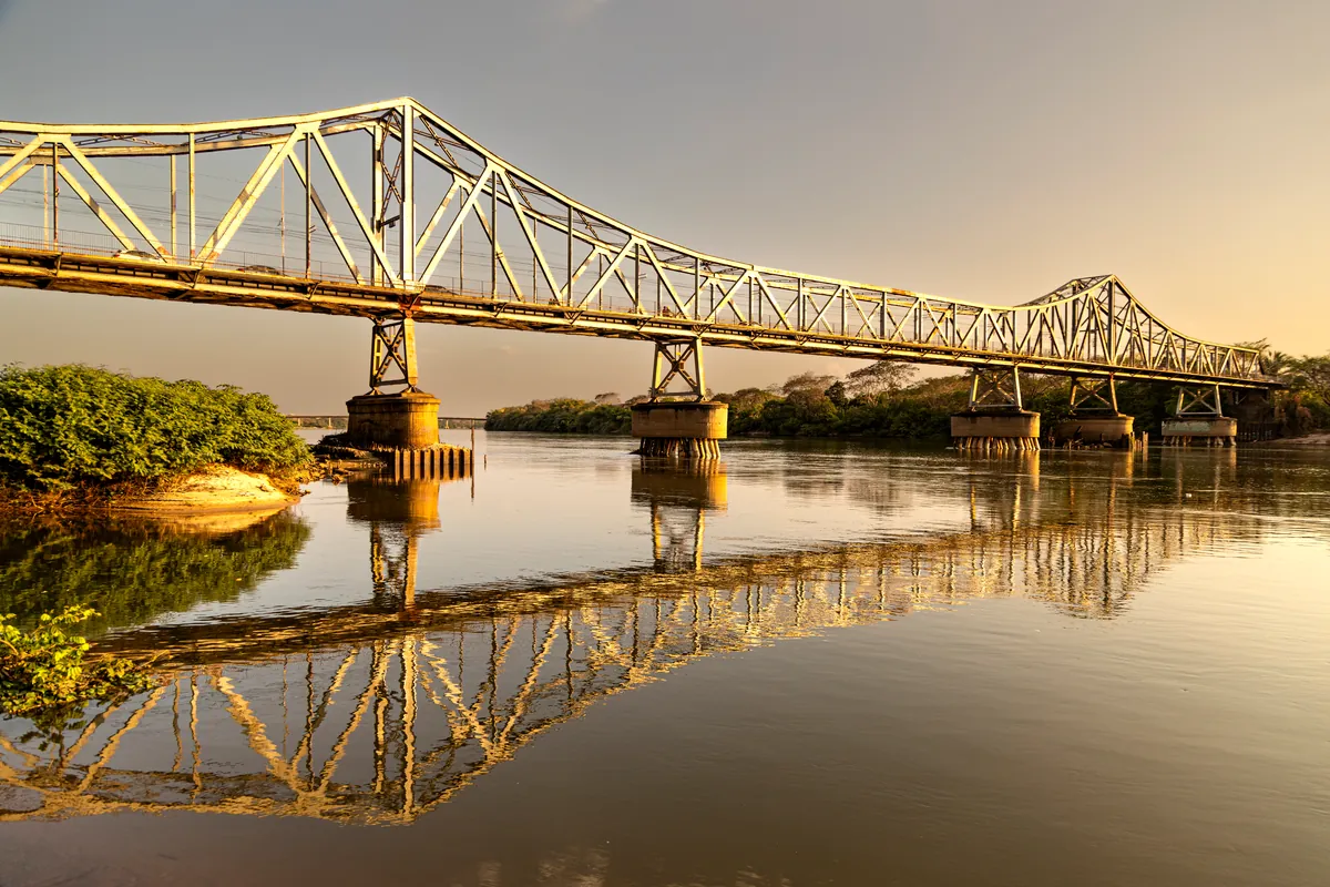 Uma ponte metálica treliçada e seu reflexo na água ao entardecer.