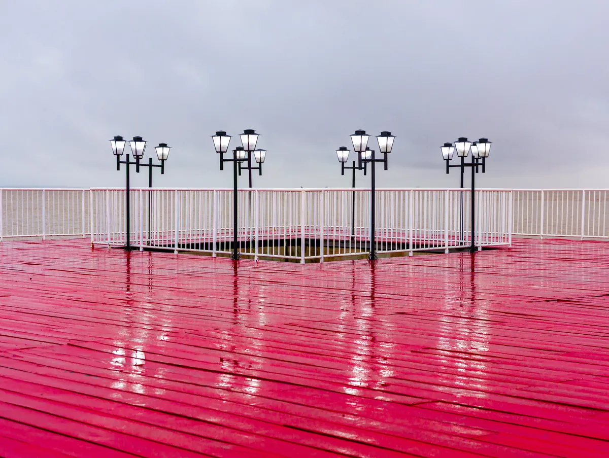 O piso vermelho molhado reflete as luminárias do trapiche sob o céu nublado.