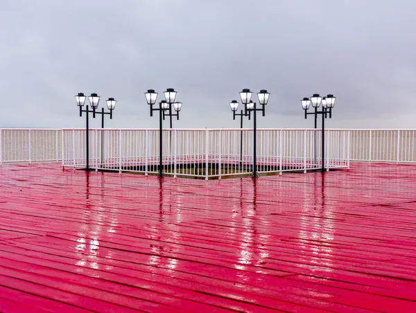 O piso vermelho molhado reflete as luminárias do trapiche sob o céu nublado.